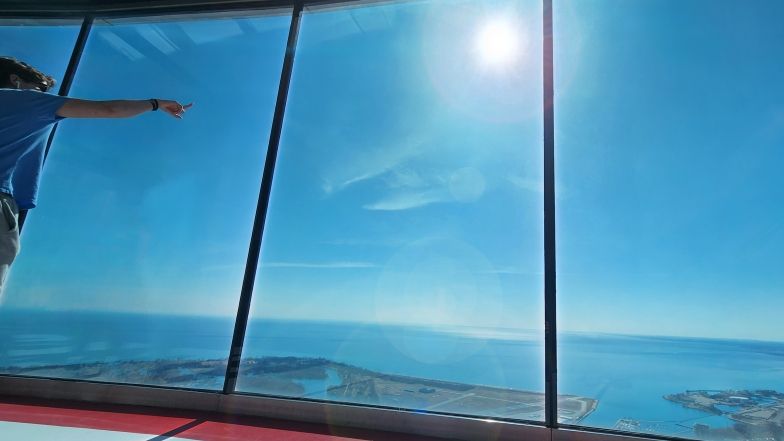 A person stands on the Main Observation Level at the CN Tower and through the floor to ceiling windows, points at the sun
