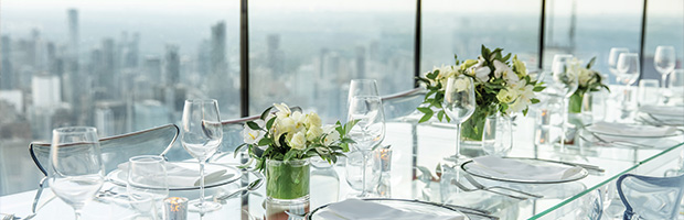 A long, clear glass table on the main observation level of the Tower, set with glassware and white floral centrepieces. Behind the table, the city skyline is visible through the windows