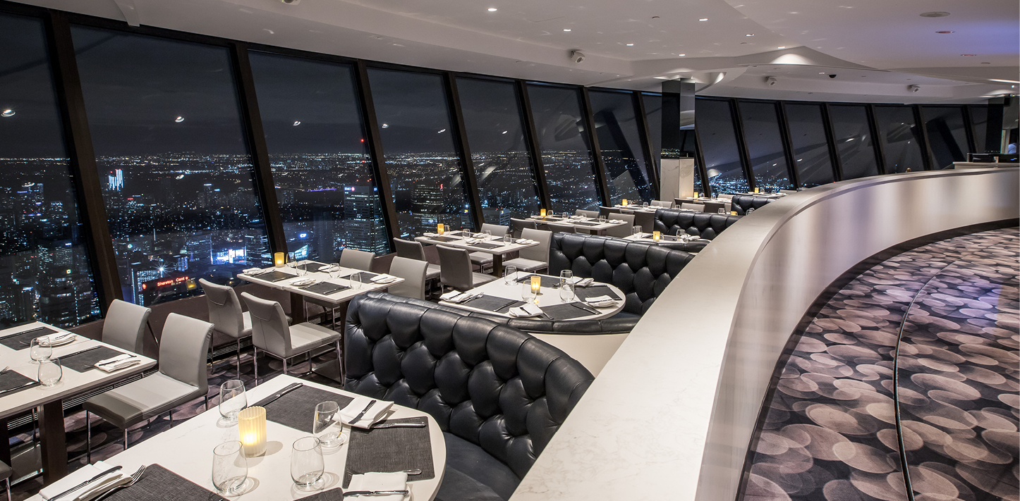 The restaurant with tables and chairs along the window and booths on the other side of the aisle. The view is overseeing Toronto at night