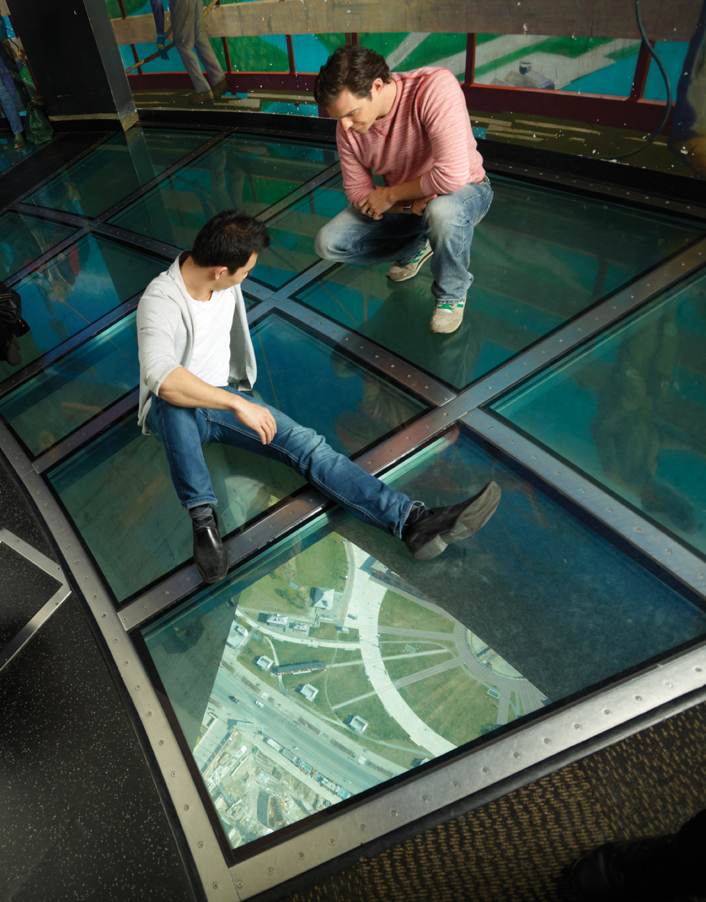 Two people sitting on the glass floor. The ground is visible far below.