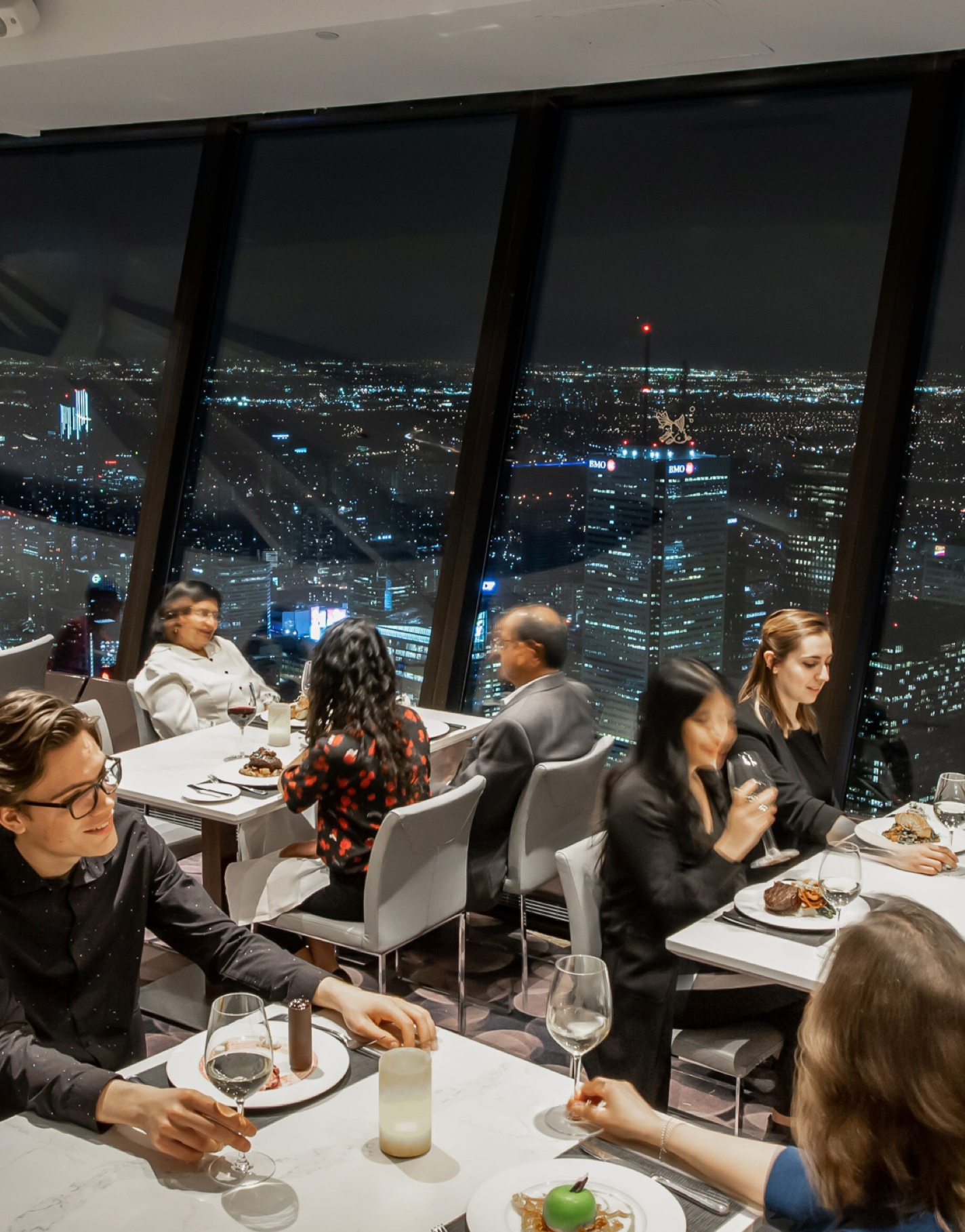 6 people sitting at 3 tables eating dinner. Toronto city lights at night in the background
