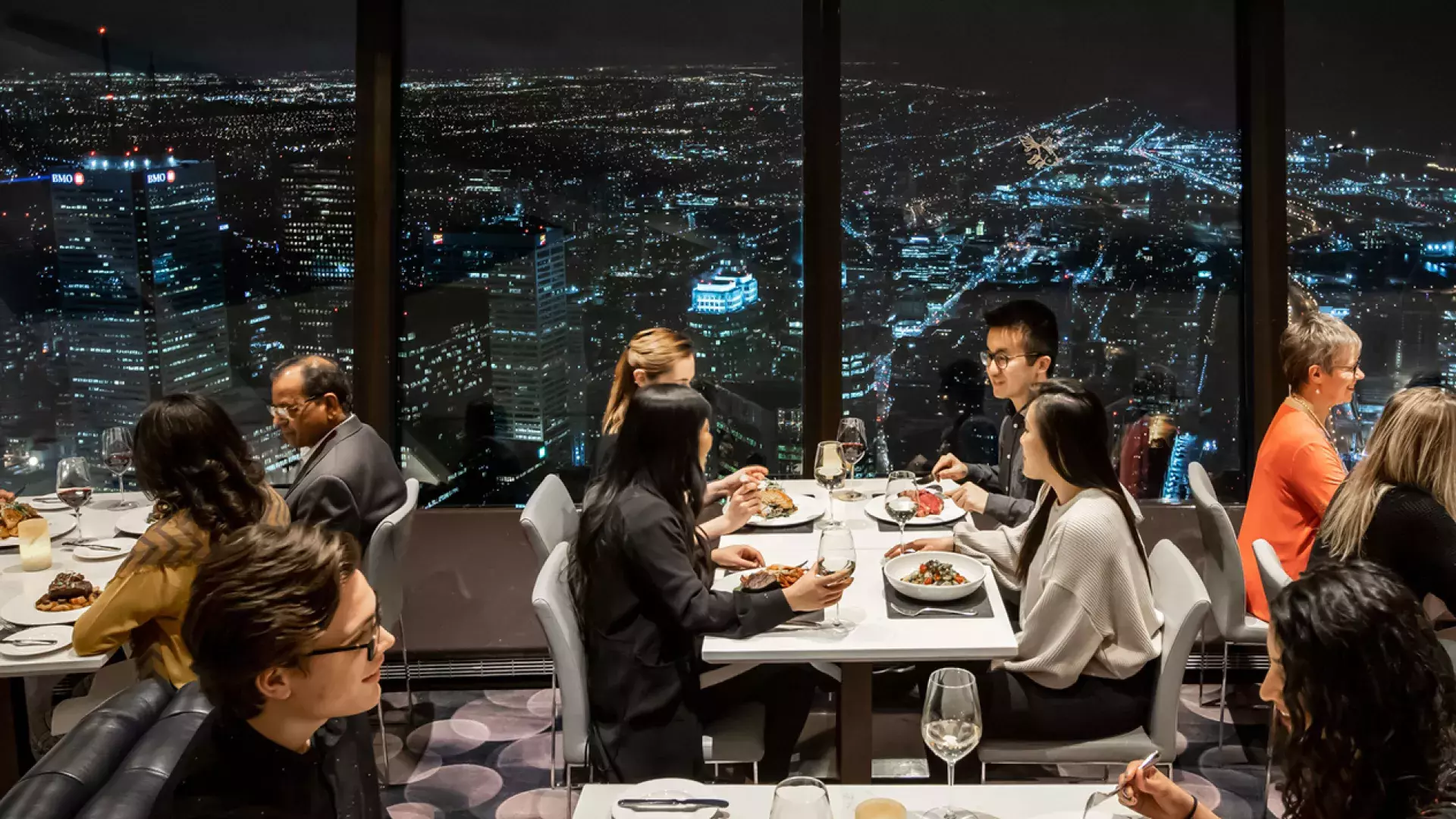 Several people seated at dining tables with the night time city lights in the background