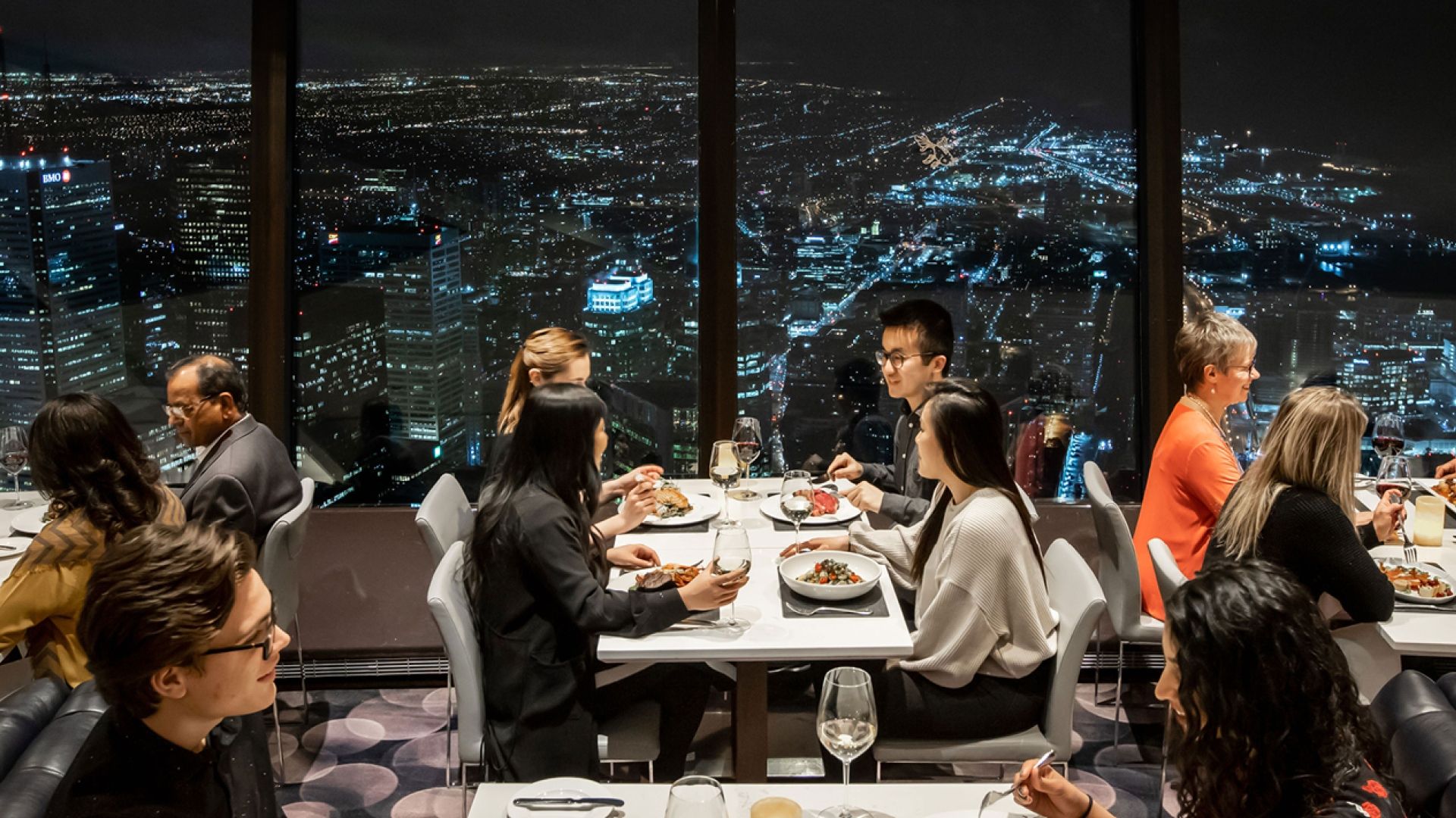 Several people seated at dining tables with the night time city lights in the background