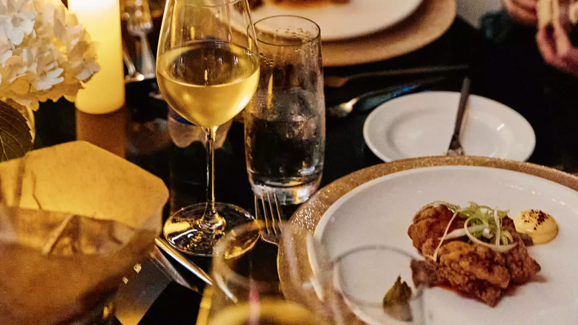 Closeup of multiple items (plate of food, glass of wine, small side plate, cutlery, flowers) dispersed on a table in the evening, lighted by a candle.