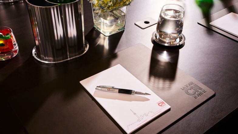 Detailed view of the meeting table with a CN Tower branded placemat, pad of paper and pen, an individual ice bucket with beverage, an empty glass, a small vase with flowers, a glass filled with candy, and a desk grommet for cord management.