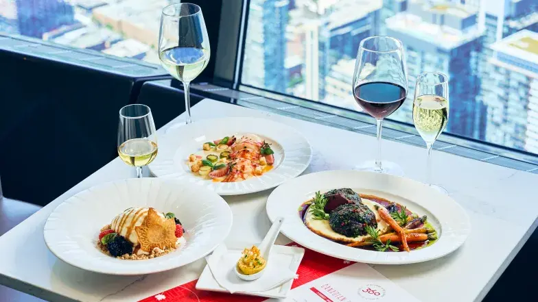 The Canadian Experience tasting menu at 360 Restaurant at the CN Tower, featuring a multi-course spread of Canadian-inspired dishes including seafood, braised meat with seasonal vegetables, dessert, and an amuse-bouche, paired with red, white, and sparkling wines. The table is set beside floor-to-ceiling windows overlooking Toronto’s downtown skyline from above.