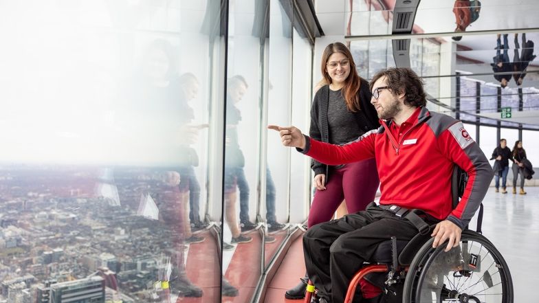 A CN Tower team member, a white man, wearing a red shirt and using a wheelchair, points something out through the floor-to-ceiling window to a guest, a white woman, standing behind him on the Main Observation Level.