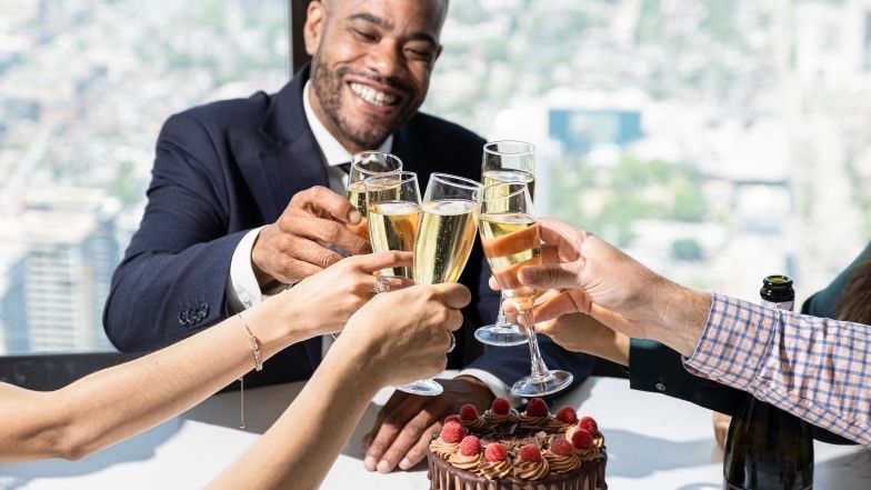 Black man toasting bubbly with friends at 360, with the view of Toronto in the back and a chocolate cake on the table.