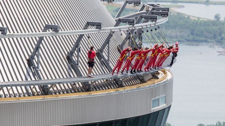 A group of people on the outside of the tower (edgewalk). They are wearing harnesses connected to security lines as they lean forward over the edge of the tower with arms straight out.