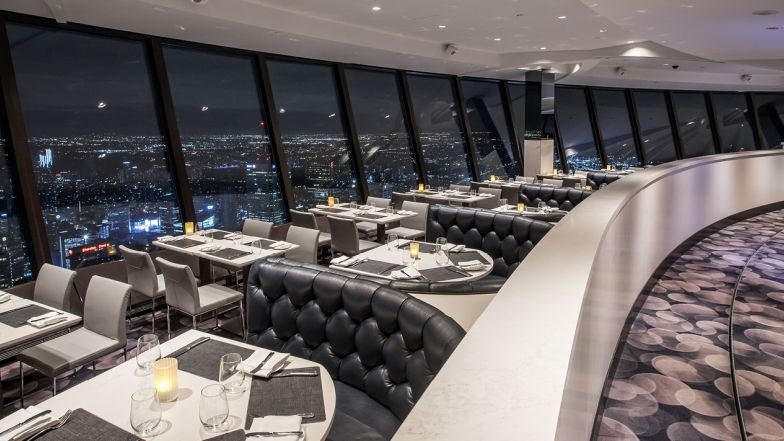 The restaurant with tables and chairs along the window and booths on the other side of the aisle. The view is overseeing Toronto at night