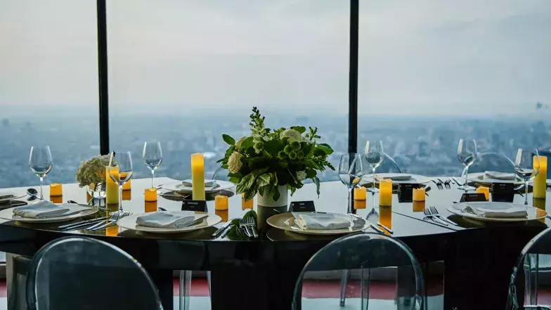 View of a serpentine table set up at the Main Observation Level of the CN Tower. On the table, we can see multiple bouquets of white flowers, surrounded by empty plates, candles and cutlery. In the background, we see the golden hour in downtown Toronto from the floor to ceiling windows.