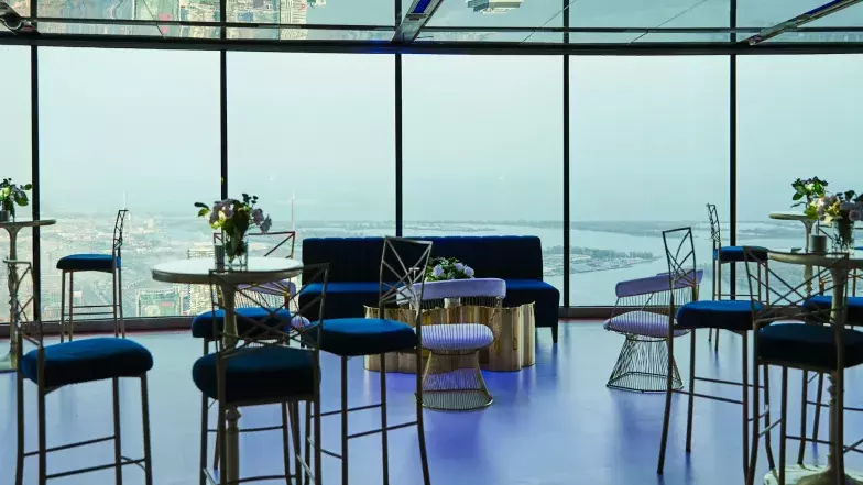 Private event setup at the Main Observation Level of the CN Tower with high top tables, couches, lamps and flowers. In the background, we see the view of Toronto and Lake Ontario through the windows.