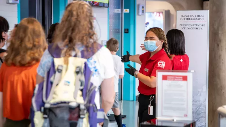 An CN Tower Employee wearing a face mask and a pair of gloves greeting people as they wait for the elevators