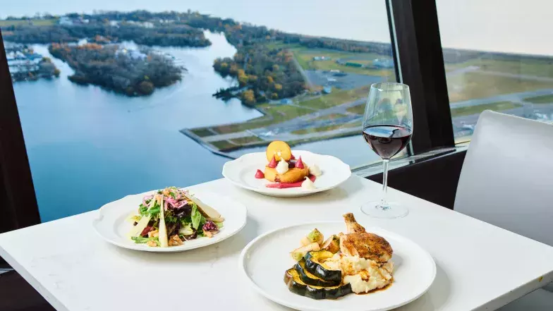 View of 3 plates of food on a white marble table at 360 restaurant. In the background, a glass of red wine and the windows of the restaurant overlooking Lake Ontario.