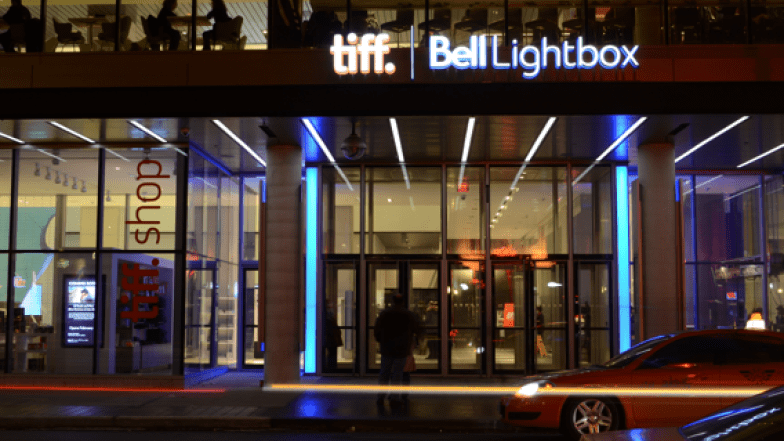 Glass wall with glass doors. Two cars passing by and a person standing in front of the building. Light sign above the doors read tiff. Bell Lightbox