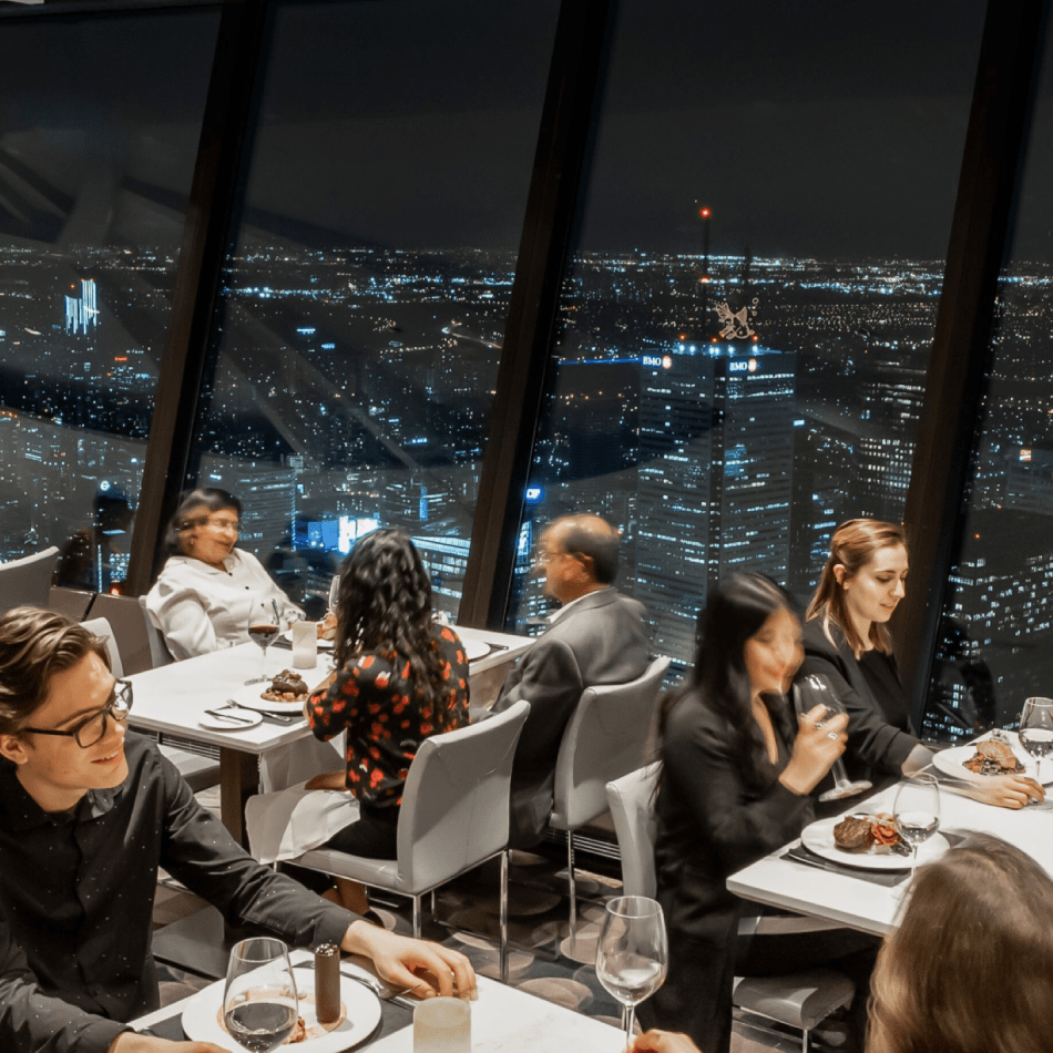 6 people sitting at 3 tables eating dinner. Toronto city lights at night in the background