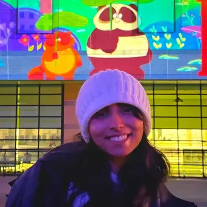 Headshot of Fatimah I outdoors at night, wearing a knit hat and jacket, with a brightly coloured mural in the background.