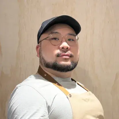 Headshot of Jin Ke Wang facing the camera indoors, wearing glasses, a cap, and an apron, against a light wood-panel background.