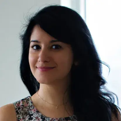 Headshot of Reena Devani facing the camera indoors, with long dark hair and a patterned top, photographed against a softly lit neutral background.
