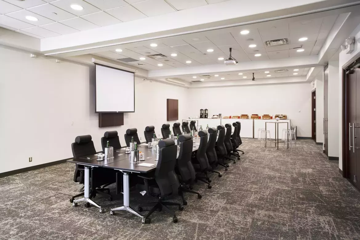 Large room with a long rectangular meeting table in the centre. The table has 8 executive chairs on each side. On the table in front of each chair is table setup. Arranged along the centre of the table are small vases with flowers and eight glasses filled with candy. On the wall next to the table and opposite the door is a pull-down projector screen. Along the far wall is a long table with food and beverages. In front of this are two square high-top tables each with four stools. 