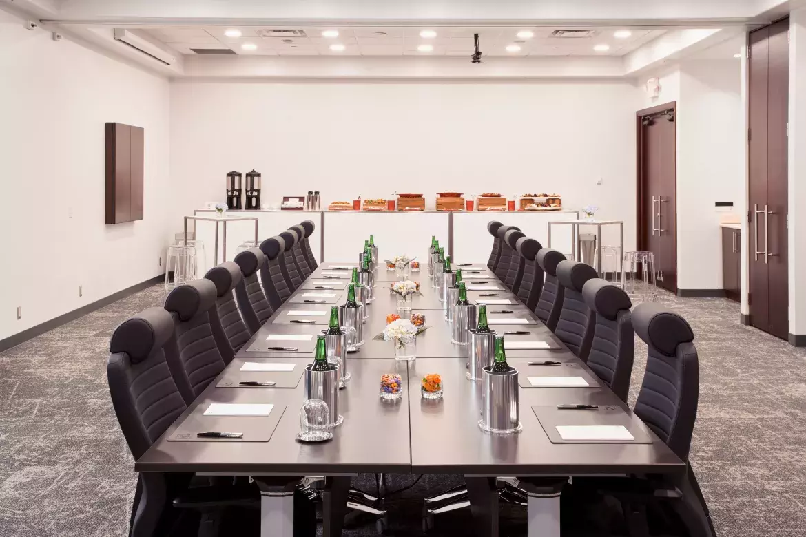 View along the centre of the meeting table towards the food and beverage table, showing small vases with flowers and glasses filled with candy, as well as placemats with pads of paper and pens, and individual ice buckets with a beverage and an empty glass at each place. 