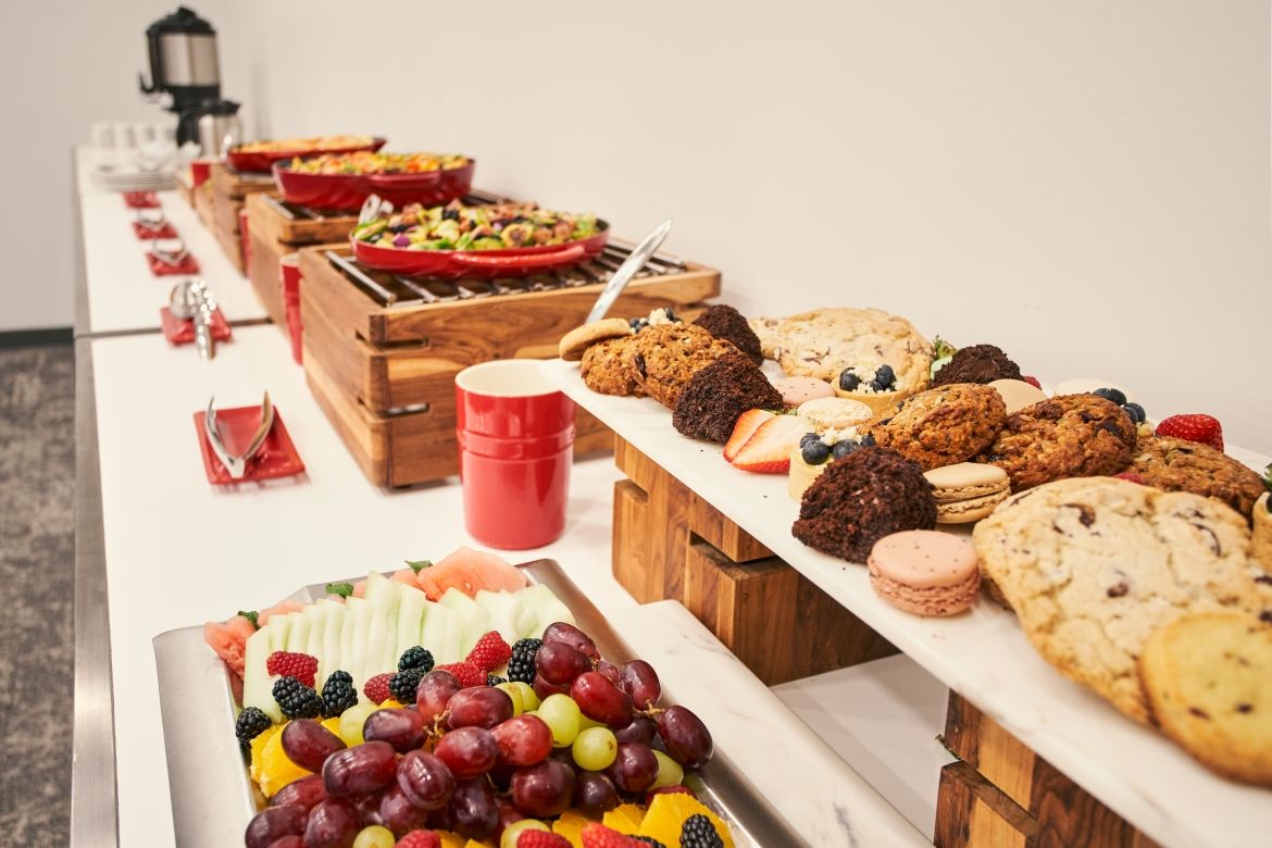 Detailed view of the other end of the food table. Dishes are presented on a rectangular marble slab raised on wooden blocks. Dishes in this view are a fruit platter and assorted cookies and brownies. 