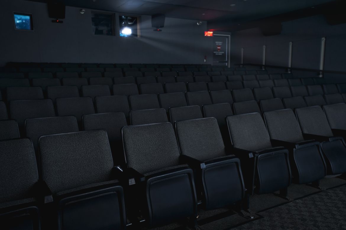 View of cinema from in front of the folded chairs. A projector window and emergency exit door can be seen in the back of the room. 