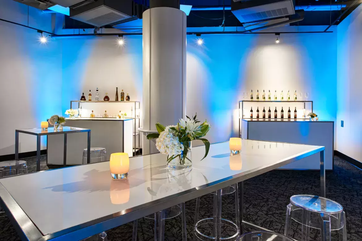 Detailed view of a rectangular high-top table with six stools in the reception area. On the table are two small candles and a vase with flowers. In the background are two bar serving areas. 