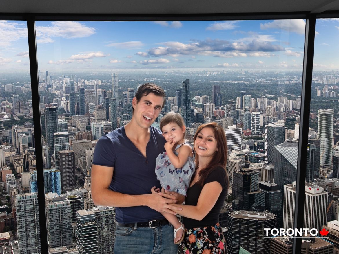 A family of three smiling for a photo in front of the cityscape view of Toronto through a large window at the CN Tower.