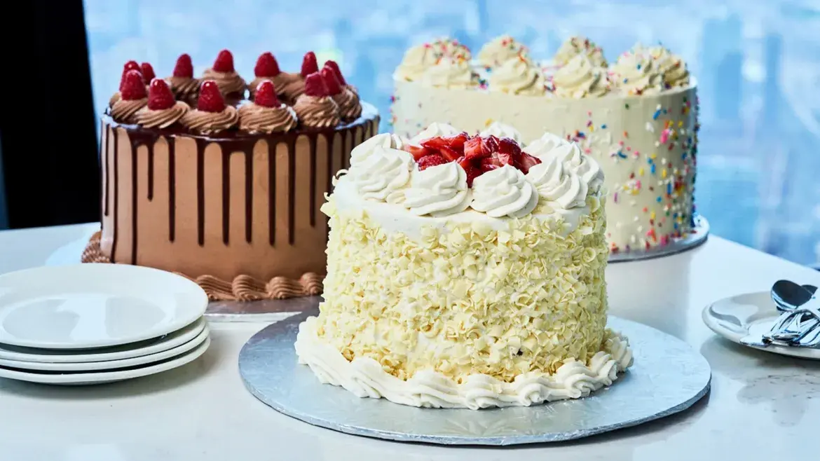 Three decorated cakes displayed on a white table at 360 Restaurant, including chocolate and vanilla cakes topped with raspberries, strawberries, frosting, and sprinkles, with dessert plates and spoons set beside angled windows overlooking the city.