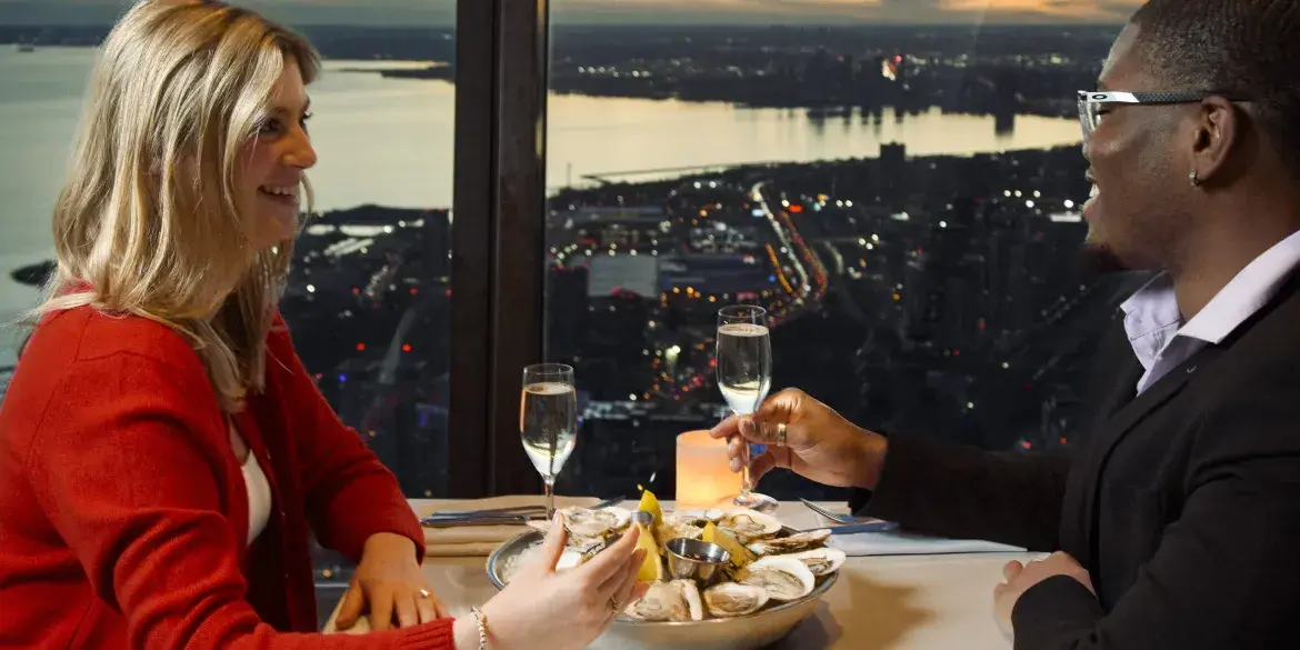 A couple enjoying fresh oysters and champagne together at 360 Restaurant. In the background, we can see the view of Toronto at night through the windows.