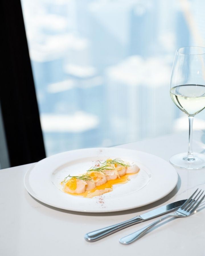 View of the scalops crudo on a white plate on a table at 360. There is also a glass of white wine and cutlery on the table. Background is an overview of the city.
